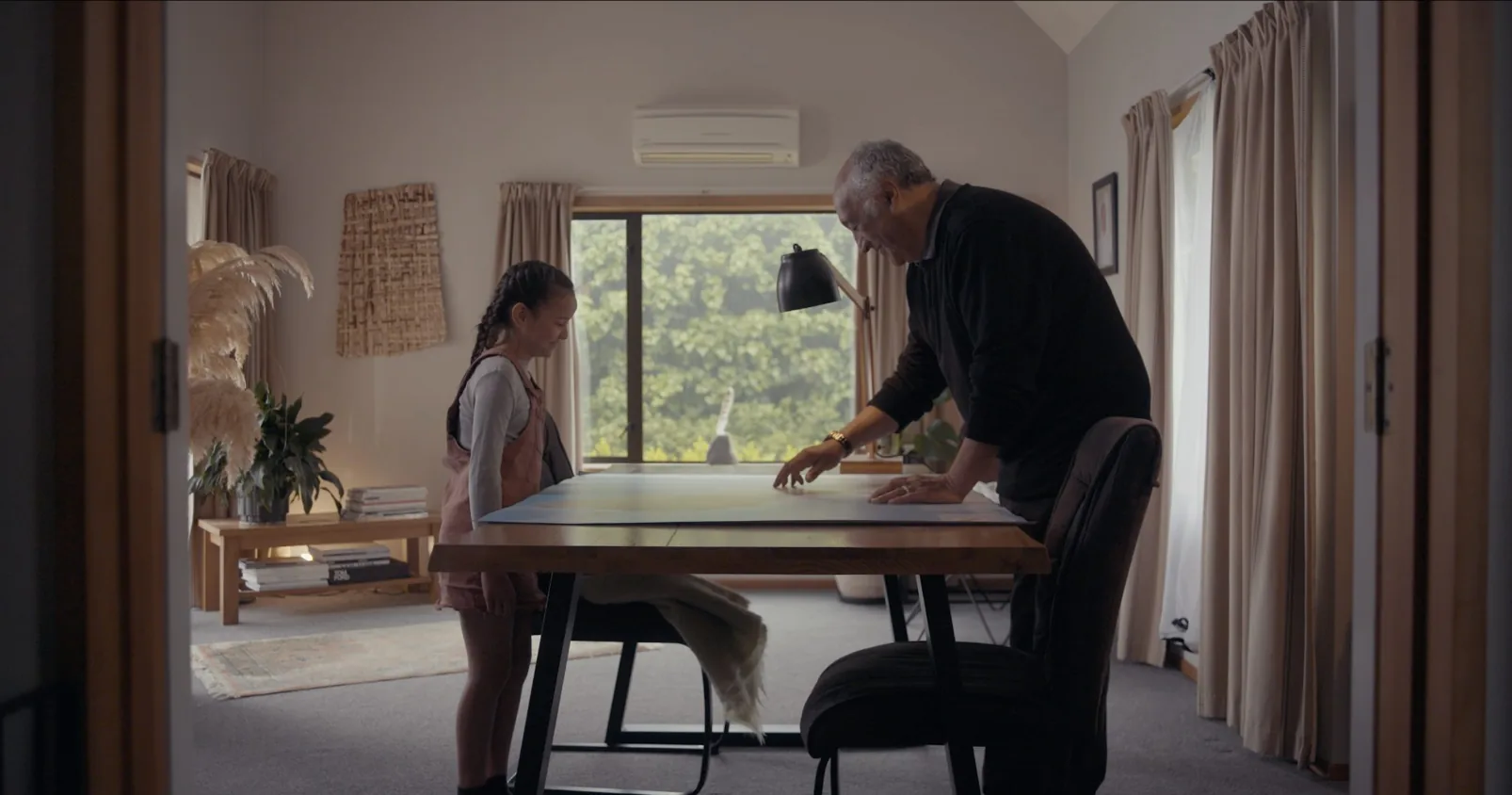 Kauraka E Mataku, Kia Takatū - Hine and Pōua leaning over a map at the dining table, soft natural light from the window behind them