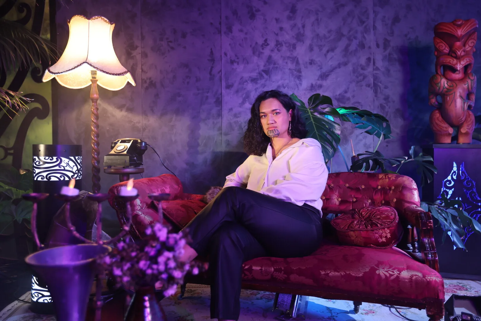 Lost in the Q - moko-clad figure reclining on a crimson chaise in a violet-lit interior with carved pou and kowhaiwhai lanterns